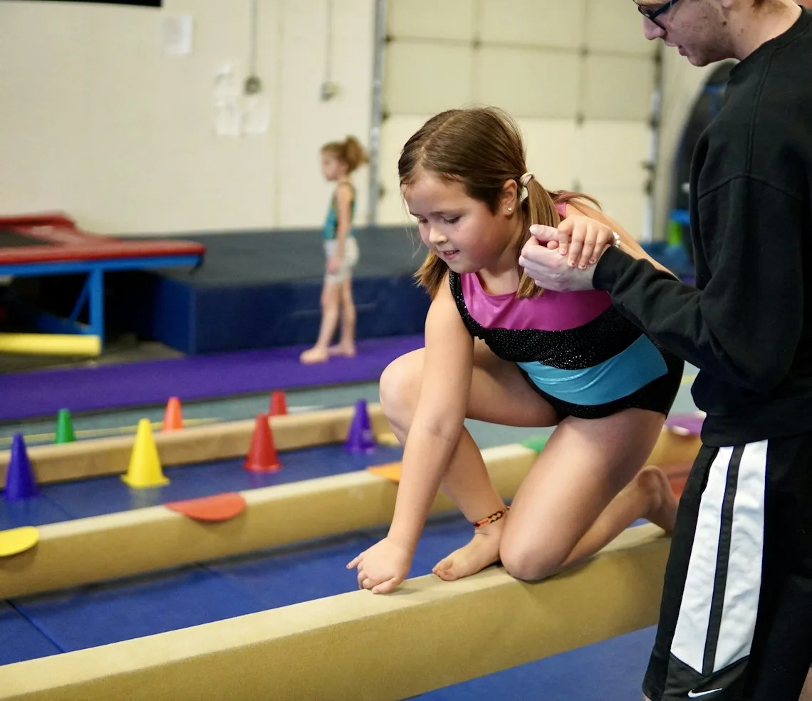 A coach spotting a girl on the balance beam in the hall
