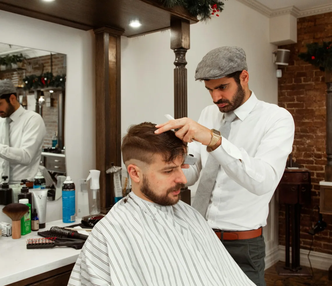 A barber in a flat cap trims a client’s hair over a comb, the client in a striped cape